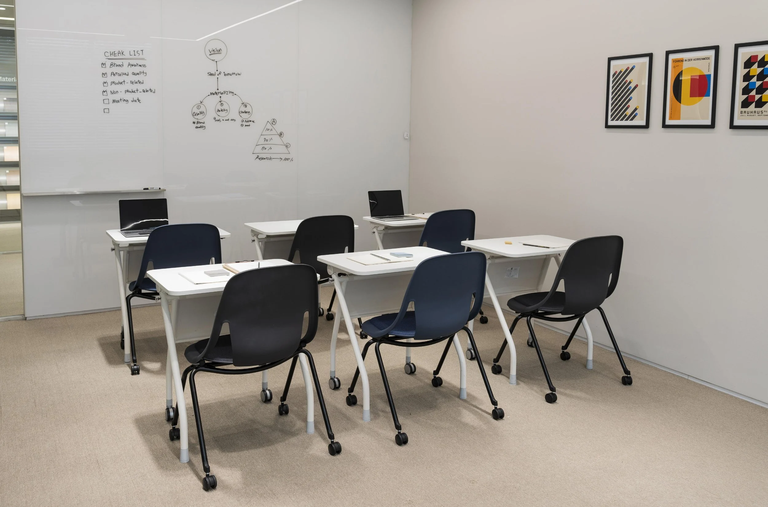 An empty classroom with white desks, black and blue chairs, a whiteboard on the left with handwritten notes, and framed abstract art on the right wall.