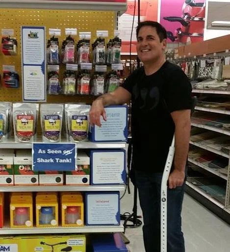 A man at a store aisle smiling and resting his arm on a display rack, with a crutch under his arm. The display has various packaged products and a sign that says "As Seen on Shark Tank!"