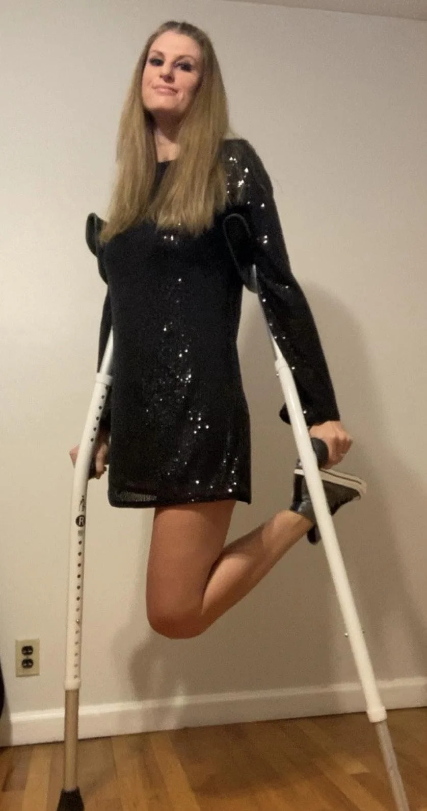 A woman with long blonde hair wearing a black sequined dress, standing on one leg, holding crutches, and looking confidently at the camera.