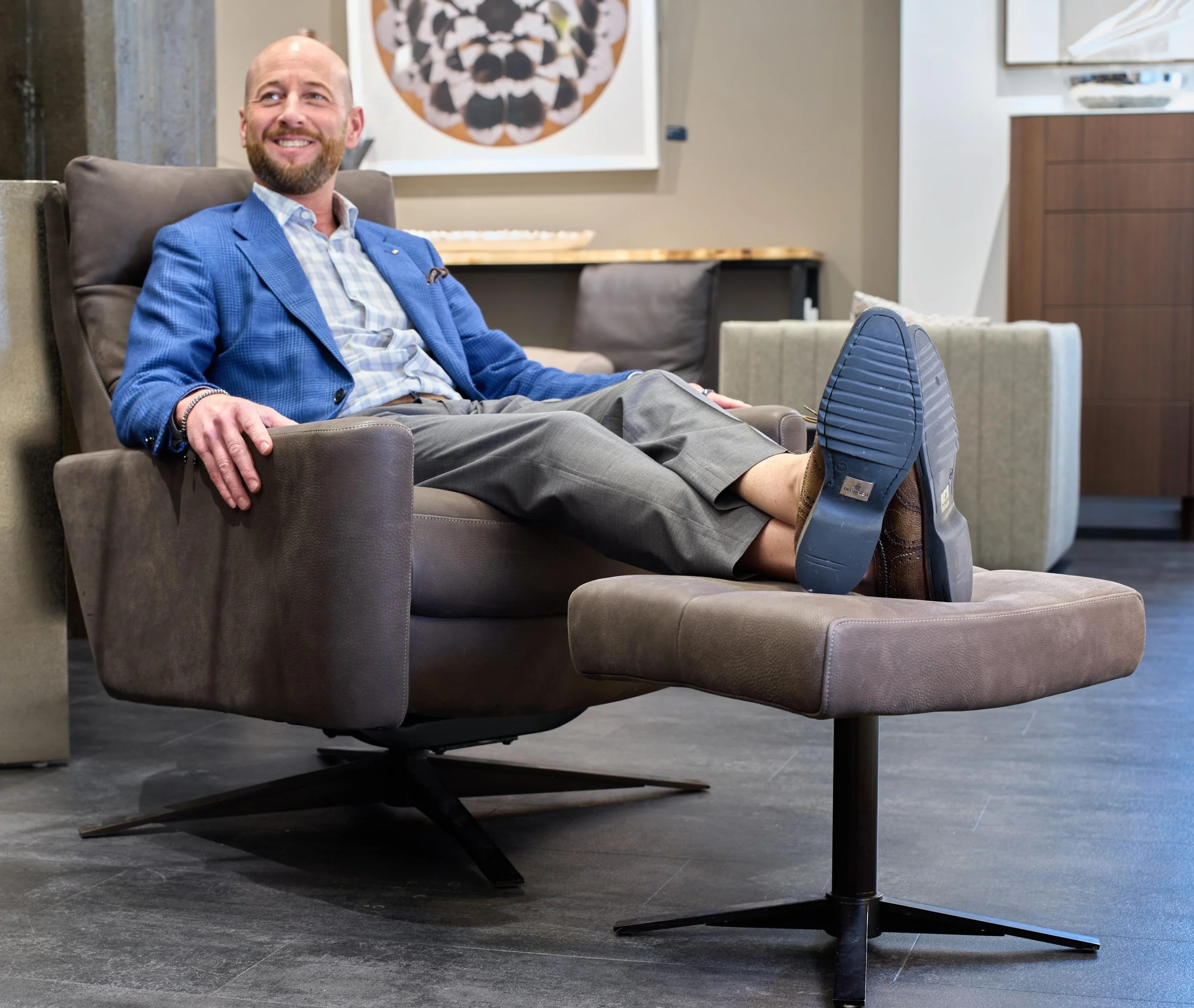 A man sitting in a leather armchair, smiling, wearing a blue blazer and gray shorts, with his feet on a round wooden table. The setting appears to be a modern art gallery or showroom with artwork hanging on the wall behind him.
