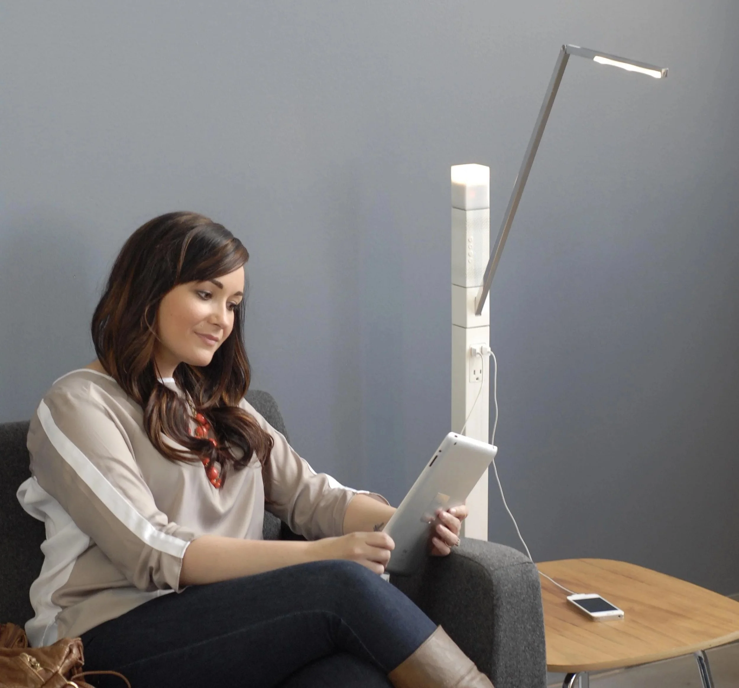 A woman with long brown hair sitting on a gray chair, looking at her smartphone, with a white lamp and a gray wall in the background.