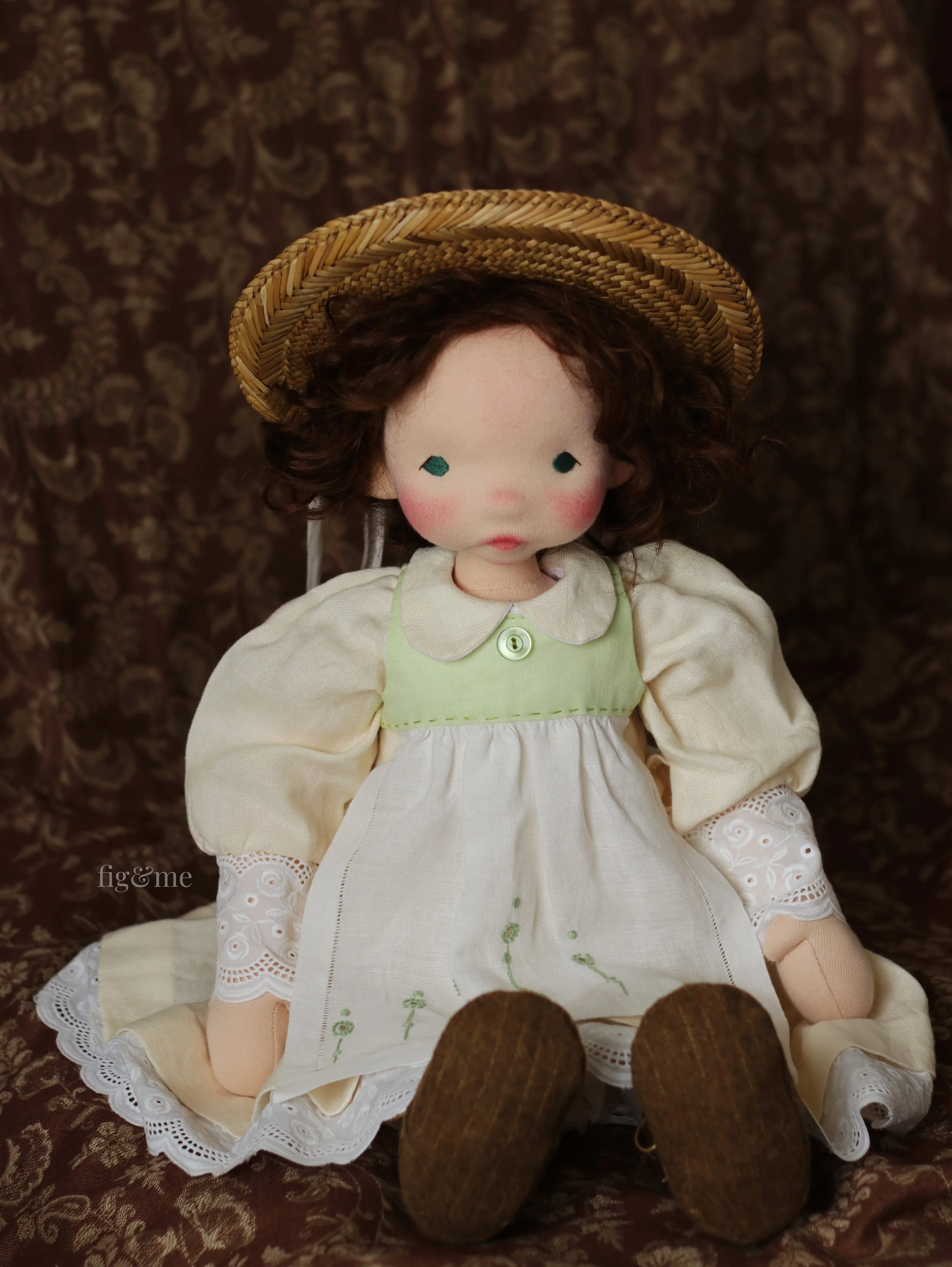 Waldorf inspired doll with brown hair, green eyes, pastel yellow linen dress and white and green pinafore, wearing an antique doll's straw hat.