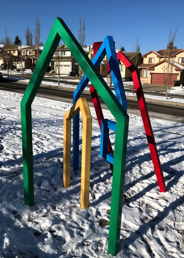 To me, from this angle all of the shapes look like houses a young child might draw. It looks very inviting for a child to run around and through it. It is also interesting to see how the shape of the artwork’s element echo those of the houses in the background.