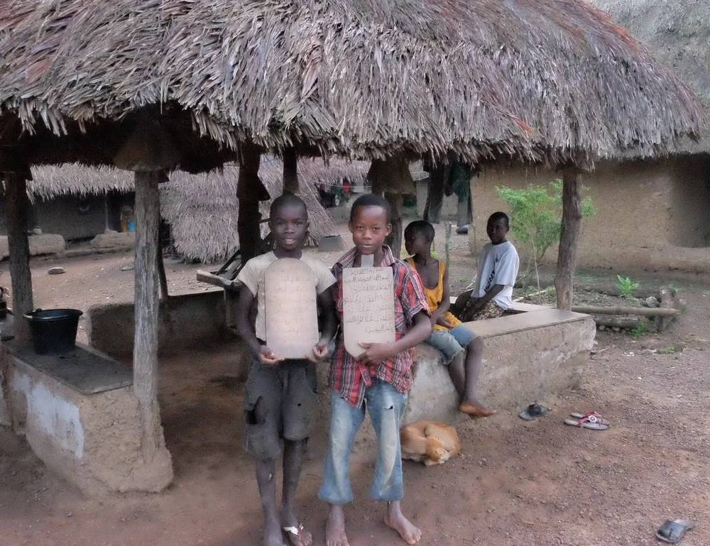 Due to a mechanical breakdown on our truck, we spent nearly an entire day in the tiny village of Kambama. The people seem very accepting of us wandering around the village and taking pictures of their houses, school, and their activities. Most of the village is Christian and their children attend the small village school. These two boys were part of the Muslim minority and they receive instruction from an Imam who visits the village 1 or 2 days each week. They were proud to display their homework neatly written in Arabic.