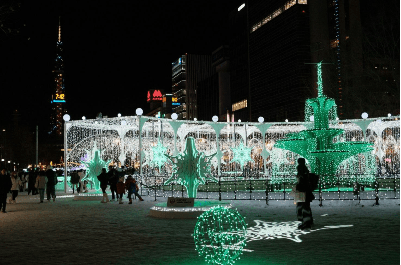 Holiday light display featuring a fountain-shaped structure, decorative snowflakes, and glowing globes in an urban city square at night, with tall buildings and a Kremlin-like tower in the background.