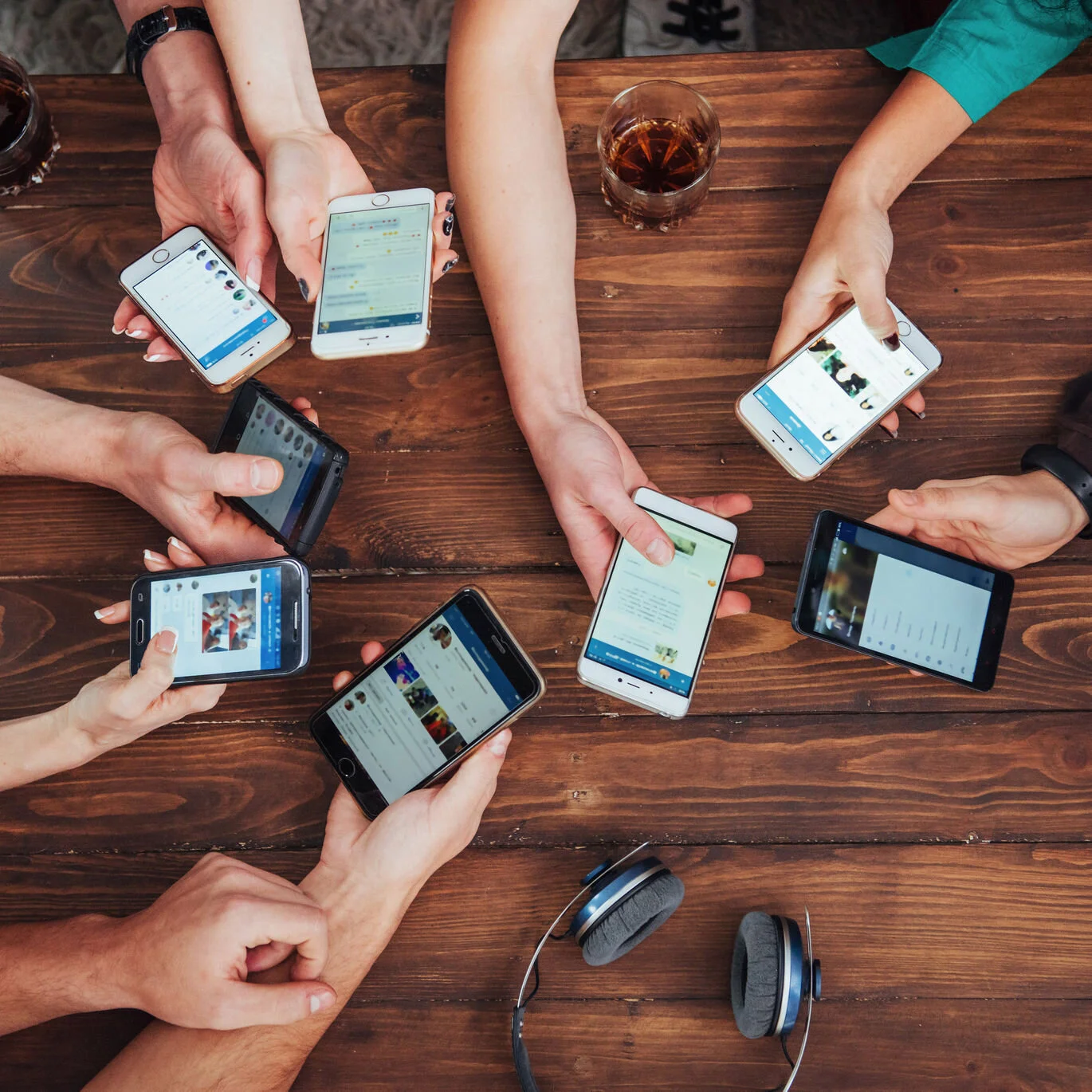 Multiple people holding smartphones around a wooden table with a glass of dark soda and a pair of headphones on it.