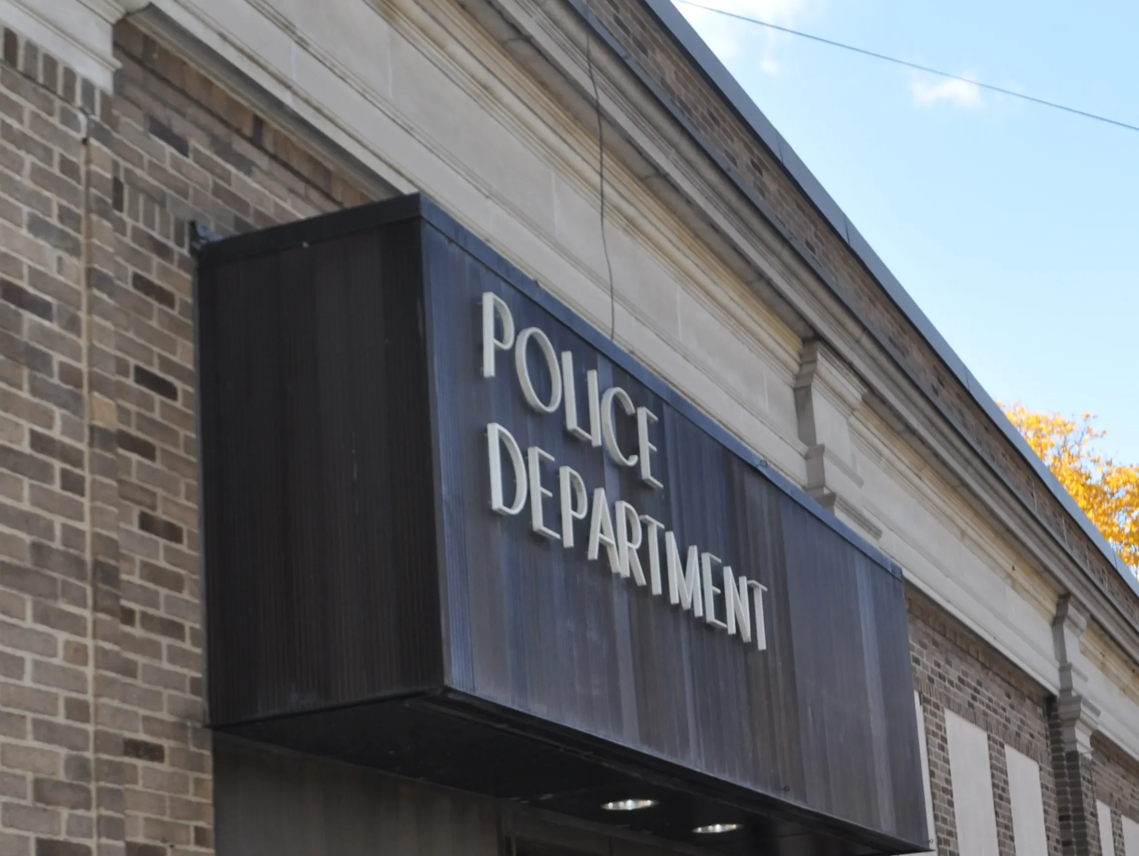 Close-up of a brick building with a black sign displaying partially visible white letters reading 'POLICE DEPARTMENT' under a blue sky.