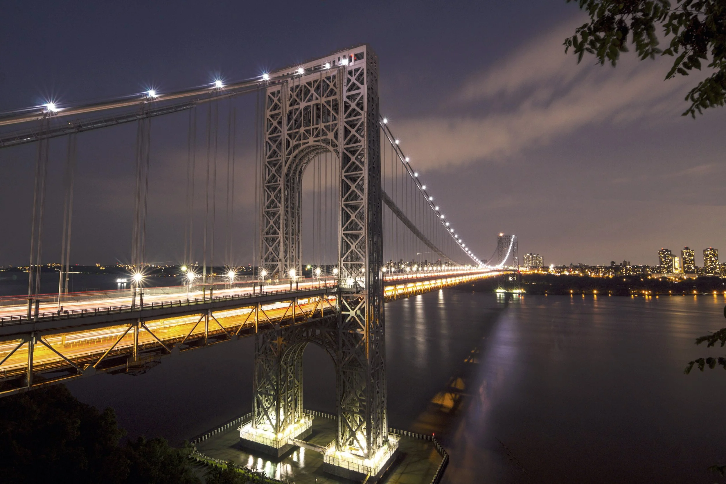 Nighttime view of the Brooklyn Bridge over the East River in New York City, illuminated with lights, with a city skyline in the background.