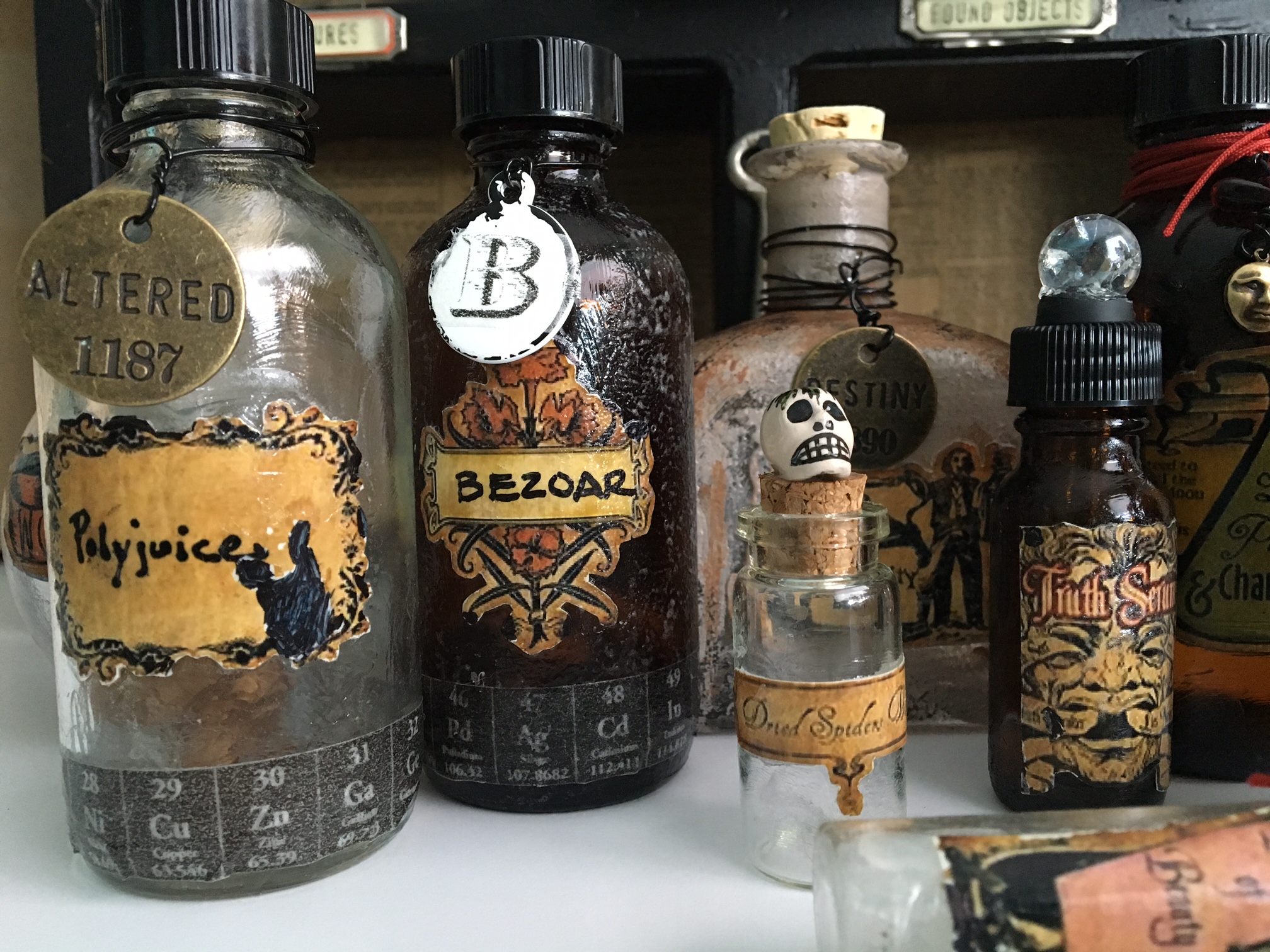 More bottle close ups. Happy accident on the "Polyjuice" bottle. The label when I was decoupaging had lifted the image leaving white. It was too big to leave, so I started coloring in with a black marker. The image started to look like a cat (which if you know the story about Hermione, you'll understand how cool that is!). I liked the metal "altered" tag, too. That's a Peruvian skull bead on the dried spider vial and a cracked marble on the truth serum.