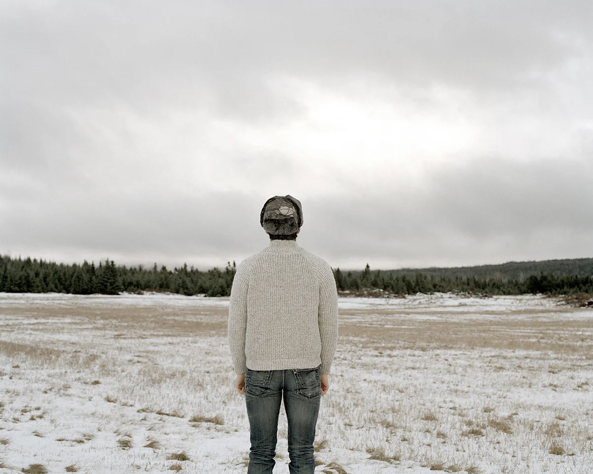 In Sublime , Adam contemplates the grandeur of the sky, ocean, and forest of his homeland in Newfoundland. As a city dweller, Adam’s experience of this expansive place is at once foreign and familiar, both about returning home and experiencing unexplored places. Sublime proved to be transformational in Adam’s work. This inaugural exploration of Newfoundland prompted further investigation of its history, leading to several bodies of work photographing resettled communities. The large scale and the focus on natural settings in these images recall 19th century Hudson River School landscape paintings. While those paintings romanticized the natural world and emphasized its formidable power over humankind, the photographs in Sublime approach nature on a less intimidating—though still majestic—scale. Newfoundland’s trademark fog is the protagonist of these photographs. Though often associated with melancholy, here the atmosphere is almost playful: clouds echo the shapes of rock formations, fog obscures the horizon line, and the only evidence of the elusive wind is the way the trees lean in its wake. The photographs are alive with texture: snow-laden branches, grassy hills, faceted cliffs, the rippled surface of the sea. Others are more nuanced, requiring a longer look, to see what lies beyond the fog. Adam inserts the human figure into some photographs in the series to investigate humankind’s relationship to the land. While figures in Hudson River School painting were minuscule compared to the imposing landscape, the figures in the Sublime photographs—seen from behind—fit into the landscape almost seamlessly. Even their clothing seems felicitous: the pattern of the woman’s sweater nearly aligns with the horizon; the man, with his camouflage hat, shares the palette of his surroundings.