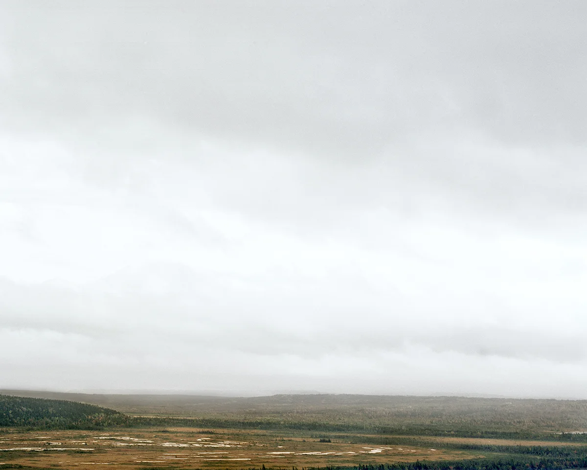 In Sublime , Adam contemplates the grandeur of the sky, ocean, and forest of his homeland in Newfoundland. As a city dweller, Adam’s experience of this expansive place is at once foreign and familiar, both about returning home and experiencing unexplored places. Sublime proved to be transformational in Adam’s work. This inaugural exploration of Newfoundland prompted further investigation of its history, leading to several bodies of work photographing resettled communities. The large scale and the focus on natural settings in these images recall 19th century Hudson River School landscape paintings. While those paintings romanticized the natural world and emphasized its formidable power over humankind, the photographs in Sublime approach nature on a less intimidating—though still majestic—scale. Newfoundland’s trademark fog is the protagonist of these photographs. Though often associated with melancholy, here the atmosphere is almost playful: clouds echo the shapes of rock formations, fog obscures the horizon line, and the only evidence of the elusive wind is the way the trees lean in its wake. The photographs are alive with texture: snow-laden branches, grassy hills, faceted cliffs, the rippled surface of the sea. Others are more nuanced, requiring a longer look, to see what lies beyond the fog. Adam inserts the human figure into some photographs in the series to investigate humankind’s relationship to the land. While figures in Hudson River School painting were minuscule compared to the imposing landscape, the figures in the Sublime photographs—seen from behind—fit into the landscape almost seamlessly. Even their clothing seems felicitous: the pattern of the woman’s sweater nearly aligns with the horizon; the man, with his camouflage hat, shares the palette of his surroundings.