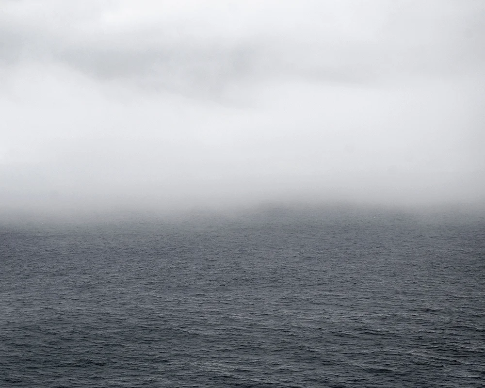 In Sublime , Adam contemplates the grandeur of the sky, ocean, and forest of his homeland in Newfoundland. As a city dweller, Adam’s experience of this expansive place is at once foreign and familiar, both about returning home and experiencing unexplored places. Sublime proved to be transformational in Adam’s work. This inaugural exploration of Newfoundland prompted further investigation of its history, leading to several bodies of work photographing resettled communities. The large scale and the focus on natural settings in these images recall 19th century Hudson River School landscape paintings. While those paintings romanticized the natural world and emphasized its formidable power over humankind, the photographs in Sublime approach nature on a less intimidating—though still majestic—scale. Newfoundland’s trademark fog is the protagonist of these photographs. Though often associated with melancholy, here the atmosphere is almost playful: clouds echo the shapes of rock formations, fog obscures the horizon line, and the only evidence of the elusive wind is the way the trees lean in its wake. The photographs are alive with texture: snow-laden branches, grassy hills, faceted cliffs, the rippled surface of the sea. Others are more nuanced, requiring a longer look, to see what lies beyond the fog. Adam inserts the human figure into some photographs in the series to investigate humankind’s relationship to the land. While figures in Hudson River School painting were minuscule compared to the imposing landscape, the figures in the Sublime photographs—seen from behind—fit into the landscape almost seamlessly. Even their clothing seems felicitous: the pattern of the woman’s sweater nearly aligns with the horizon; the man, with his camouflage hat, shares the palette of his surroundings.