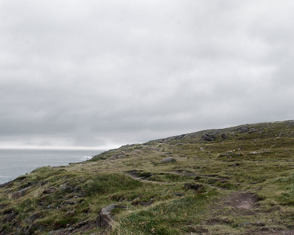 In Sublime , Adam contemplates the grandeur of the sky, ocean, and forest of his homeland in Newfoundland. As a city dweller, Adam’s experience of this expansive place is at once foreign and familiar, both about returning home and experiencing unexplored places. Sublime proved to be transformational in Adam’s work. This inaugural exploration of Newfoundland prompted further investigation of its history, leading to several bodies of work photographing resettled communities. The large scale and the focus on natural settings in these images recall 19th century Hudson River School landscape paintings. While those paintings romanticized the natural world and emphasized its formidable power over humankind, the photographs in Sublime approach nature on a less intimidating—though still majestic—scale. Newfoundland’s trademark fog is the protagonist of these photographs. Though often associated with melancholy, here the atmosphere is almost playful: clouds echo the shapes of rock formations, fog obscures the horizon line, and the only evidence of the elusive wind is the way the trees lean in its wake. The photographs are alive with texture: snow-laden branches, grassy hills, faceted cliffs, the rippled surface of the sea. Others are more nuanced, requiring a longer look, to see what lies beyond the fog. Adam inserts the human figure into some photographs in the series to investigate humankind’s relationship to the land. While figures in Hudson River School painting were minuscule compared to the imposing landscape, the figures in the Sublime photographs—seen from behind—fit into the landscape almost seamlessly. Even their clothing seems felicitous: the pattern of the woman’s sweater nearly aligns with the horizon; the man, with his camouflage hat, shares the palette of his surroundings.