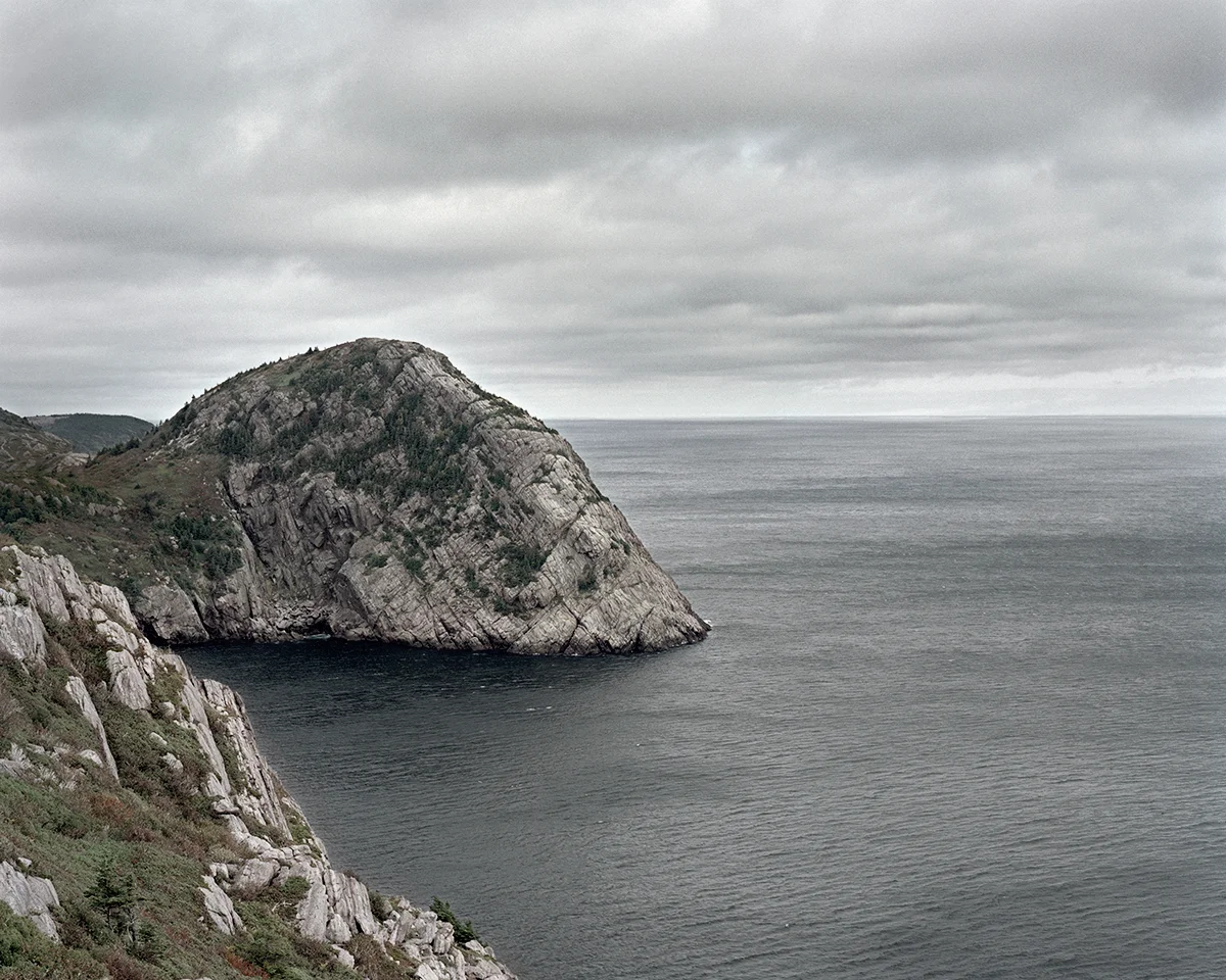 In Sublime , Adam contemplates the grandeur of the sky, ocean, and forest of his homeland in Newfoundland. As a city dweller, Adam’s experience of this expansive place is at once foreign and familiar, both about returning home and experiencing unexplored places. Sublime proved to be transformational in Adam’s work. This inaugural exploration of Newfoundland prompted further investigation of its history, leading to several bodies of work photographing resettled communities. The large scale and the focus on natural settings in these images recall 19th century Hudson River School landscape paintings. While those paintings romanticized the natural world and emphasized its formidable power over humankind, the photographs in Sublime approach nature on a less intimidating—though still majestic—scale. Newfoundland’s trademark fog is the protagonist of these photographs. Though often associated with melancholy, here the atmosphere is almost playful: clouds echo the shapes of rock formations, fog obscures the horizon line, and the only evidence of the elusive wind is the way the trees lean in its wake. The photographs are alive with texture: snow-laden branches, grassy hills, faceted cliffs, the rippled surface of the sea. Others are more nuanced, requiring a longer look, to see what lies beyond the fog. Adam inserts the human figure into some photographs in the series to investigate humankind’s relationship to the land. While figures in Hudson River School painting were minuscule compared to the imposing landscape, the figures in the Sublime photographs—seen from behind—fit into the landscape almost seamlessly. Even their clothing seems felicitous: the pattern of the woman’s sweater nearly aligns with the horizon; the man, with his camouflage hat, shares the palette of his surroundings.