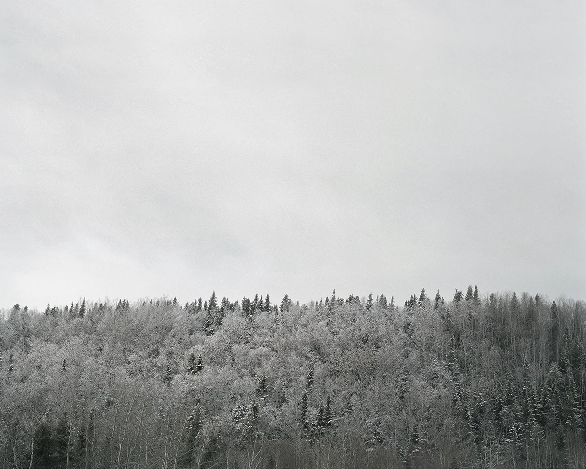 In Sublime , Adam contemplates the grandeur of the sky, ocean, and forest of his homeland in Newfoundland. As a city dweller, Adam’s experience of this expansive place is at once foreign and familiar, both about returning home and experiencing unexplored places. Sublime proved to be transformational in Adam’s work. This inaugural exploration of Newfoundland prompted further investigation of its history, leading to several bodies of work photographing resettled communities. The large scale and the focus on natural settings in these images recall 19th century Hudson River School landscape paintings. While those paintings romanticized the natural world and emphasized its formidable power over humankind, the photographs in Sublime approach nature on a less intimidating—though still majestic—scale. Newfoundland’s trademark fog is the protagonist of these photographs. Though often associated with melancholy, here the atmosphere is almost playful: clouds echo the shapes of rock formations, fog obscures the horizon line, and the only evidence of the elusive wind is the way the trees lean in its wake. The photographs are alive with texture: snow-laden branches, grassy hills, faceted cliffs, the rippled surface of the sea. Others are more nuanced, requiring a longer look, to see what lies beyond the fog. Adam inserts the human figure into some photographs in the series to investigate humankind’s relationship to the land. While figures in Hudson River School painting were minuscule compared to the imposing landscape, the figures in the Sublime photographs—seen from behind—fit into the landscape almost seamlessly. Even their clothing seems felicitous: the pattern of the woman’s sweater nearly aligns with the horizon; the man, with his camouflage hat, shares the palette of his surroundings.