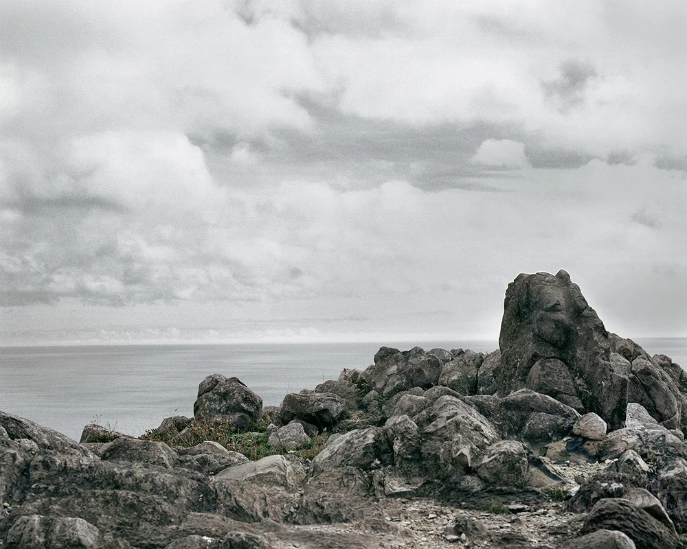 In Sublime , Adam contemplates the grandeur of the sky, ocean, and forest of his homeland in Newfoundland. As a city dweller, Adam’s experience of this expansive place is at once foreign and familiar, both about returning home and experiencing unexplored places. Sublime proved to be transformational in Adam’s work. This inaugural exploration of Newfoundland prompted further investigation of its history, leading to several bodies of work photographing resettled communities. The large scale and the focus on natural settings in these images recall 19th century Hudson River School landscape paintings. While those paintings romanticized the natural world and emphasized its formidable power over humankind, the photographs in Sublime approach nature on a less intimidating—though still majestic—scale. Newfoundland’s trademark fog is the protagonist of these photographs. Though often associated with melancholy, here the atmosphere is almost playful: clouds echo the shapes of rock formations, fog obscures the horizon line, and the only evidence of the elusive wind is the way the trees lean in its wake. The photographs are alive with texture: snow-laden branches, grassy hills, faceted cliffs, the rippled surface of the sea. Others are more nuanced, requiring a longer look, to see what lies beyond the fog. Adam inserts the human figure into some photographs in the series to investigate humankind’s relationship to the land. While figures in Hudson River School painting were minuscule compared to the imposing landscape, the figures in the Sublime photographs—seen from behind—fit into the landscape almost seamlessly. Even their clothing seems felicitous: the pattern of the woman’s sweater nearly aligns with the horizon; the man, with his camouflage hat, shares the palette of his surroundings.