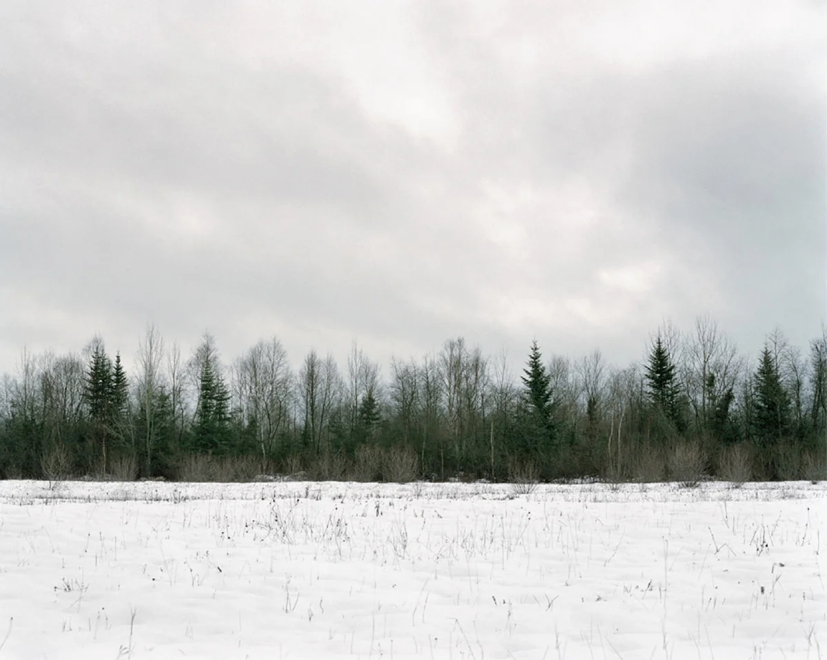 In Sublime , Adam contemplates the grandeur of the sky, ocean, and forest of his homeland in Newfoundland. As a city dweller, Adam’s experience of this expansive place is at once foreign and familiar, both about returning home and experiencing unexplored places. Sublime proved to be transformational in Adam’s work. This inaugural exploration of Newfoundland prompted further investigation of its history, leading to several bodies of work photographing resettled communities. The large scale and the focus on natural settings in these images recall 19th century Hudson River School landscape paintings. While those paintings romanticized the natural world and emphasized its formidable power over humankind, the photographs in Sublime approach nature on a less intimidating—though still majestic—scale. Newfoundland’s trademark fog is the protagonist of these photographs. Though often associated with melancholy, here the atmosphere is almost playful: clouds echo the shapes of rock formations, fog obscures the horizon line, and the only evidence of the elusive wind is the way the trees lean in its wake. The photographs are alive with texture: snow-laden branches, grassy hills, faceted cliffs, the rippled surface of the sea. Others are more nuanced, requiring a longer look, to see what lies beyond the fog. Adam inserts the human figure into some photographs in the series to investigate humankind’s relationship to the land. While figures in Hudson River School painting were minuscule compared to the imposing landscape, the figures in the Sublime photographs—seen from behind—fit into the landscape almost seamlessly. Even their clothing seems felicitous: the pattern of the woman’s sweater nearly aligns with the horizon; the man, with his camouflage hat, shares the palette of his surroundings.