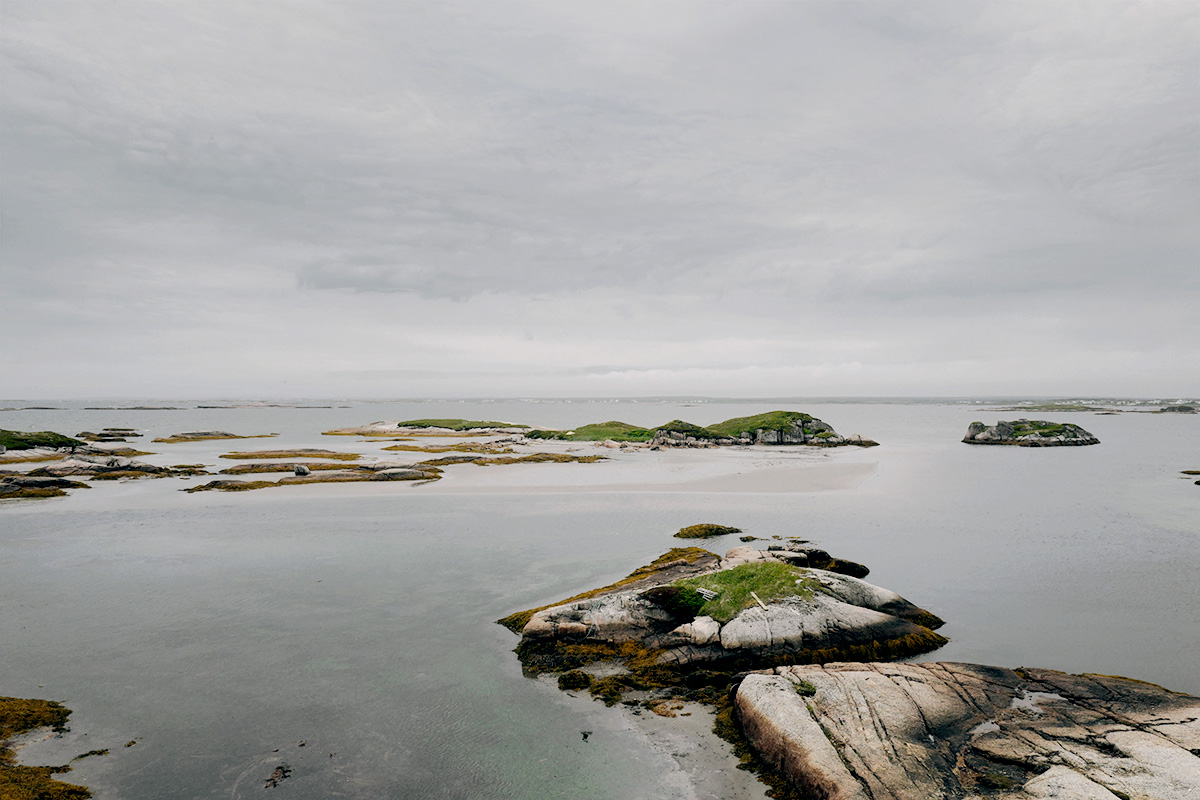 In Pinchard’s Island, Adam documents his family’s reclaiming of the land where his grandmother was born, before the Canadian government’s Resettlement Act paid residents to move from the isolated fishing community to areas with more potential for economic growth. Over a series of trips to the remote island, Adam and his family have built a cabin there, staking a claim to the land that has been all but forgotten, its people dispersed, and its culture left to fade away. Through this hyper-local and deeply personal series of large-scale photographs, Adam evokes global themes of displacement, homogenizing of cultures, and the inextricability of place from an individual’s—and family’s—identity. The photographs in this series highlight the contrast between the desolate landscape and the animation that the reestablishment of even the smallest community brings to the setting. One element Adam manipulates to create this contrast is scale: in one photograph, the built structures are barely noticeable in the sweeping panorama of the shoreline, while in another, the interior space looms large as the windows frame and contain the expanse of the land.
