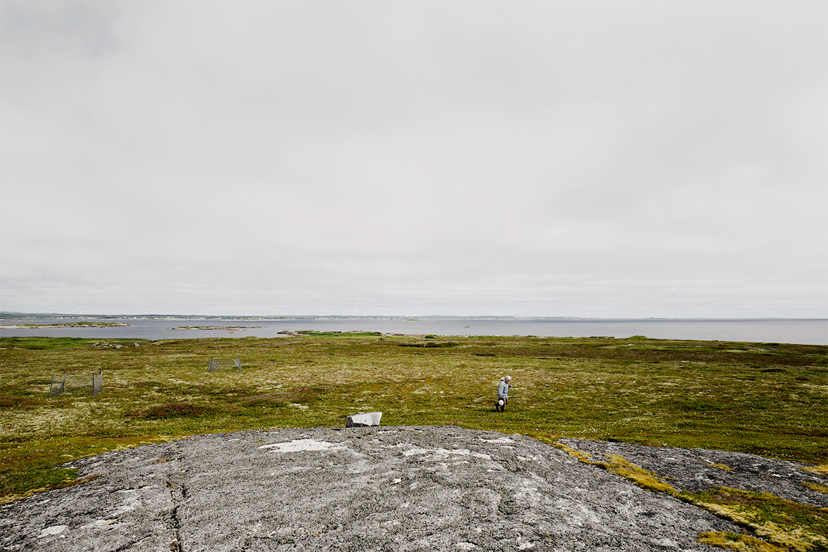In Pinchard’s Island, Adam documents his family’s reclaiming of the land where his grandmother was born, before the Canadian government’s Resettlement Act paid residents to move from the isolated fishing community to areas with more potential for economic growth. Over a series of trips to the remote island, Adam and his family have built a cabin there, staking a claim to the land that has been all but forgotten, its people dispersed, and its culture left to fade away. Through this hyper-local and deeply personal series of large-scale photographs, Adam evokes global themes of displacement, homogenizing of cultures, and the inextricability of place from an individual’s—and family’s—identity. The photographs in this series highlight the contrast between the desolate landscape and the animation that the reestablishment of even the smallest community brings to the setting. One element Adam manipulates to create this contrast is scale: in one photograph, the built structures are barely noticeable in the sweeping panorama of the shoreline, while in another, the interior space looms large as the windows frame and contain the expanse of the land.