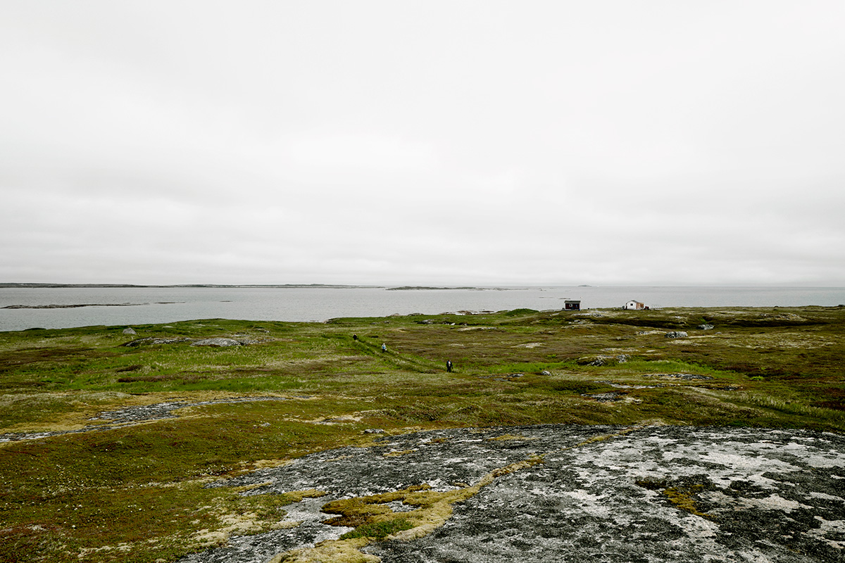 In Pinchard’s Island, Adam documents his family’s reclaiming of the land where his grandmother was born, before the Canadian government’s Resettlement Act paid residents to move from the isolated fishing community to areas with more potential for economic growth. Over a series of trips to the remote island, Adam and his family have built a cabin there, staking a claim to the land that has been all but forgotten, its people dispersed, and its culture left to fade away. Through this hyper-local and deeply personal series of large-scale photographs, Adam evokes global themes of displacement, homogenizing of cultures, and the inextricability of place from an individual’s—and family’s—identity. The photographs in this series highlight the contrast between the desolate landscape and the animation that the reestablishment of even the smallest community brings to the setting. One element Adam manipulates to create this contrast is scale: in one photograph, the built structures are barely noticeable in the sweeping panorama of the shoreline, while in another, the interior space looms large as the windows frame and contain the expanse of the land.
