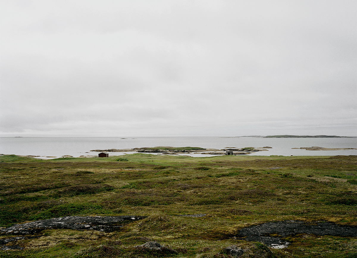 In Pinchard’s Island, Adam documents his family’s reclaiming of the land where his grandmother was born, before the Canadian government’s Resettlement Act paid residents to move from the isolated fishing community to areas with more potential for economic growth. Over a series of trips to the remote island, Adam and his family have built a cabin there, staking a claim to the land that has been all but forgotten, its people dispersed, and its culture left to fade away. Through this hyper-local and deeply personal series of large-scale photographs, Adam evokes global themes of displacement, homogenizing of cultures, and the inextricability of place from an individual’s—and family’s—identity. The photographs in this series highlight the contrast between the desolate landscape and the animation that the reestablishment of even the smallest community brings to the setting. One element Adam manipulates to create this contrast is scale: in one photograph, the built structures are barely noticeable in the sweeping panorama of the shoreline, while in another, the interior space looms large as the windows frame and contain the expanse of the land.