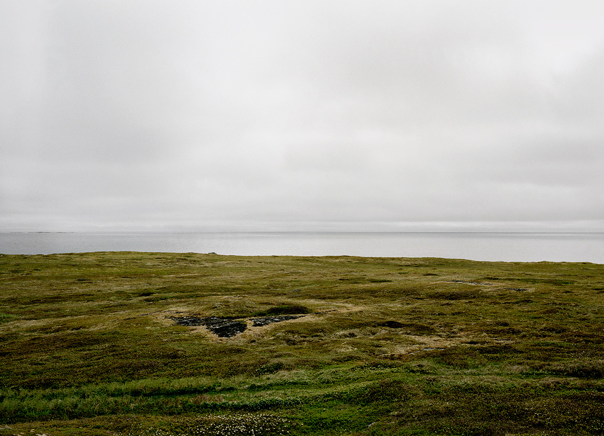 In Pinchard’s Island, Adam documents his family’s reclaiming of the land where his grandmother was born, before the Canadian government’s Resettlement Act paid residents to move from the isolated fishing community to areas with more potential for economic growth. Over a series of trips to the remote island, Adam and his family have built a cabin there, staking a claim to the land that has been all but forgotten, its people dispersed, and its culture left to fade away. Through this hyper-local and deeply personal series of large-scale photographs, Adam evokes global themes of displacement, homogenizing of cultures, and the inextricability of place from an individual’s—and family’s—identity. The photographs in this series highlight the contrast between the desolate landscape and the animation that the reestablishment of even the smallest community brings to the setting. One element Adam manipulates to create this contrast is scale: in one photograph, the built structures are barely noticeable in the sweeping panorama of the shoreline, while in another, the interior space looms large as the windows frame and contain the expanse of the land.