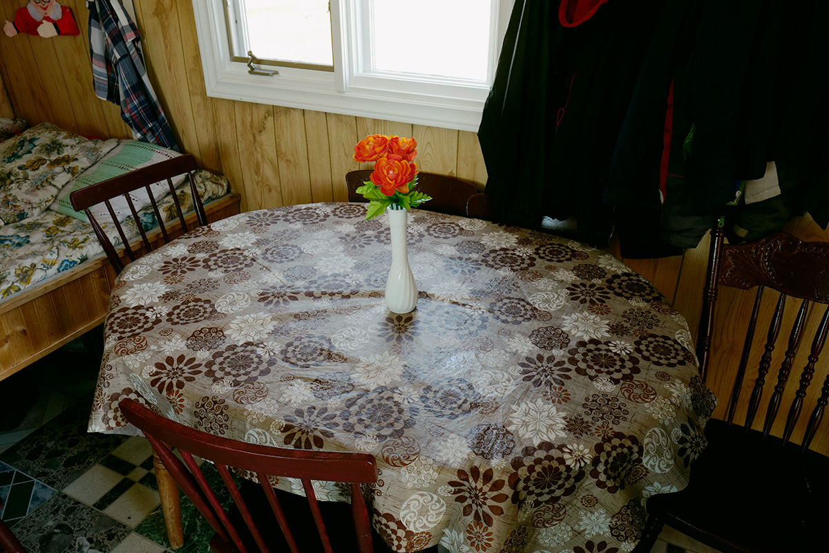 In Pinchard’s Island, Adam documents his family’s reclaiming of the land where his grandmother was born, before the Canadian government’s Resettlement Act paid residents to move from the isolated fishing community to areas with more potential for economic growth. Over a series of trips to the remote island, Adam and his family have built a cabin there, staking a claim to the land that has been all but forgotten, its people dispersed, and its culture left to fade away. Through this hyper-local and deeply personal series of large-scale photographs, Adam evokes global themes of displacement, homogenizing of cultures, and the inextricability of place from an individual’s—and family’s—identity. The photographs in this series highlight the contrast between the desolate landscape and the animation that the reestablishment of even the smallest community brings to the setting. One element Adam manipulates to create this contrast is scale: in one photograph, the built structures are barely noticeable in the sweeping panorama of the shoreline, while in another, the interior space looms large as the windows frame and contain the expanse of the land.