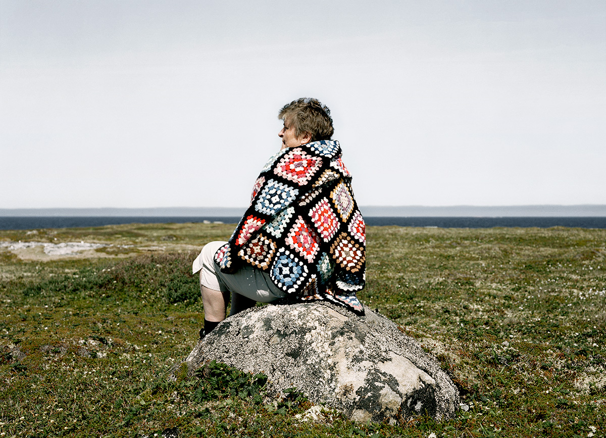 In Pinchard’s Island, Adam documents his family’s reclaiming of the land where his grandmother was born, before the Canadian government’s Resettlement Act paid residents to move from the isolated fishing community to areas with more potential for economic growth. Over a series of trips to the remote island, Adam and his family have built a cabin there, staking a claim to the land that has been all but forgotten, its people dispersed, and its culture left to fade away. Through this hyper-local and deeply personal series of large-scale photographs, Adam evokes global themes of displacement, homogenizing of cultures, and the inextricability of place from an individual’s—and family’s—identity. The photographs in this series highlight the contrast between the desolate landscape and the animation that the reestablishment of even the smallest community brings to the setting. One element Adam manipulates to create this contrast is scale: in one photograph, the built structures are barely noticeable in the sweeping panorama of the shoreline, while in another, the interior space looms large as the windows frame and contain the expanse of the land.