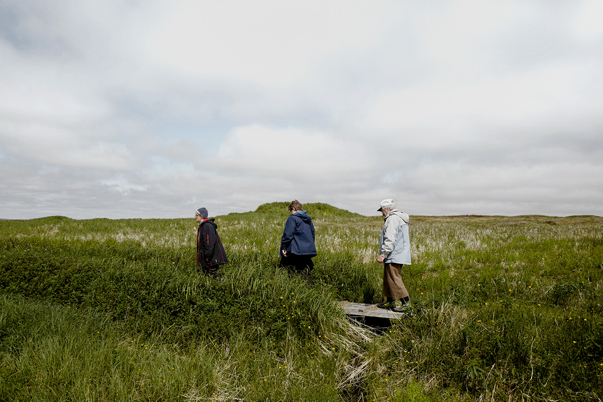 In Pinchard’s Island, Adam documents his family’s reclaiming of the land where his grandmother was born, before the Canadian government’s Resettlement Act paid residents to move from the isolated fishing community to areas with more potential for economic growth. Over a series of trips to the remote island, Adam and his family have built a cabin there, staking a claim to the land that has been all but forgotten, its people dispersed, and its culture left to fade away. Through this hyper-local and deeply personal series of large-scale photographs, Adam evokes global themes of displacement, homogenizing of cultures, and the inextricability of place from an individual’s—and family’s—identity. The photographs in this series highlight the contrast between the desolate landscape and the animation that the reestablishment of even the smallest community brings to the setting. One element Adam manipulates to create this contrast is scale: in one photograph, the built structures are barely noticeable in the sweeping panorama of the shoreline, while in another, the interior space looms large as the windows frame and contain the expanse of the land.