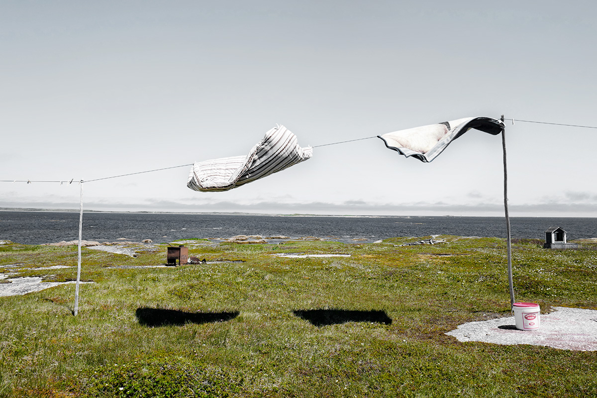 In Pinchard’s Island, Adam documents his family’s reclaiming of the land where his grandmother was born, before the Canadian government’s Resettlement Act paid residents to move from the isolated fishing community to areas with more potential for economic growth. Over a series of trips to the remote island, Adam and his family have built a cabin there, staking a claim to the land that has been all but forgotten, its people dispersed, and its culture left to fade away. Through this hyper-local and deeply personal series of large-scale photographs, Adam evokes global themes of displacement, homogenizing of cultures, and the inextricability of place from an individual’s—and family’s—identity. The photographs in this series highlight the contrast between the desolate landscape and the animation that the reestablishment of even the smallest community brings to the setting. One element Adam manipulates to create this contrast is scale: in one photograph, the built structures are barely noticeable in the sweeping panorama of the shoreline, while in another, the interior space looms large as the windows frame and contain the expanse of the land.