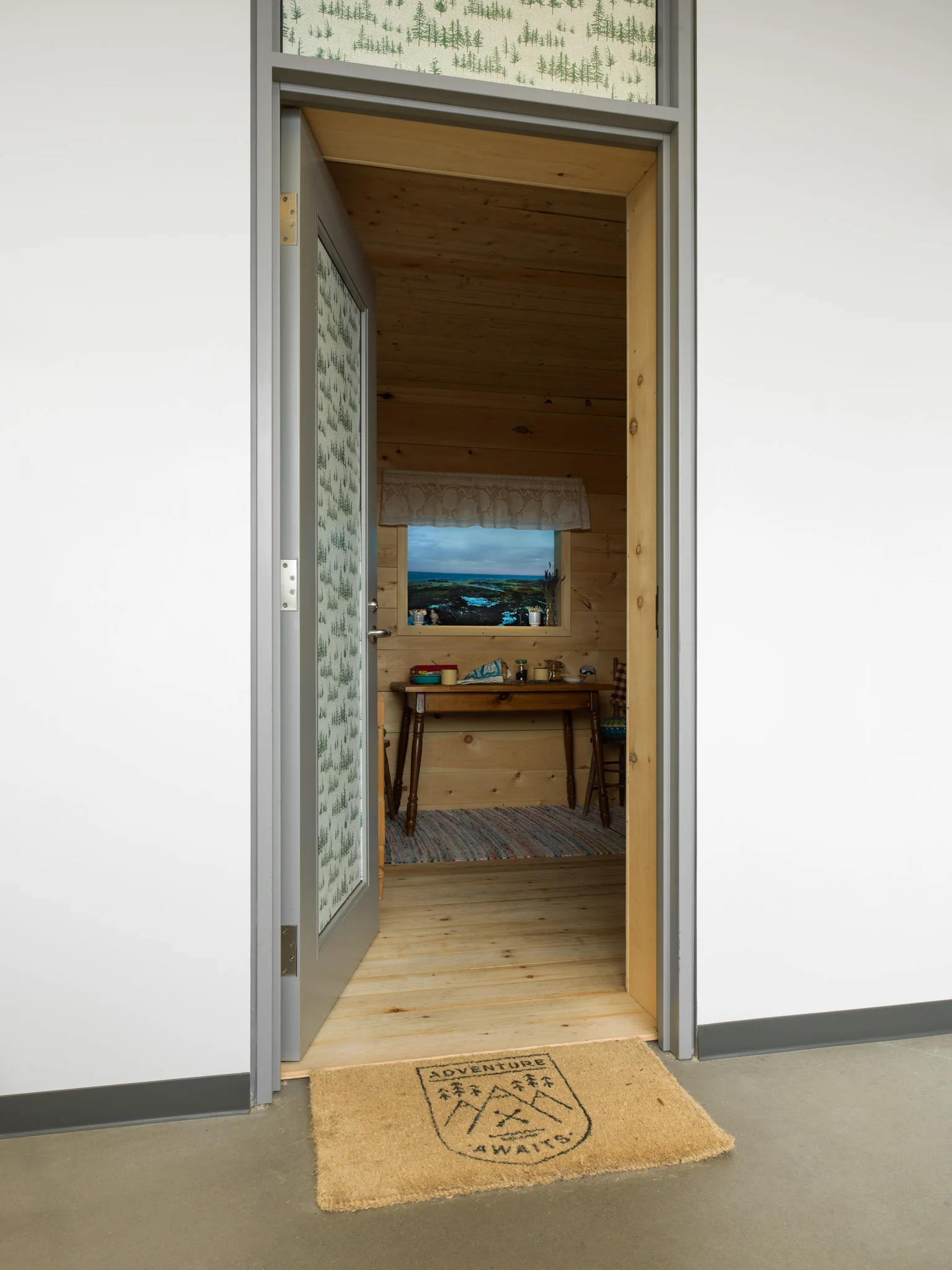 Opening a nondescript door in the corridor of a high-rise building and unexpectedly stepping into a traditional Newfoundland cabin is a disorienting experience. Indeed, entering Cabin feels like traveling back in time, or even into a completely different realm. In this immersive installation, Adam has completely transformed a 10-foot square studio space, obscuring all traces of the original structure. Visitors find themselves in the interior of a cabin that Adam built, assisted by his father, using materials sourced from the family’s homeland of Newfoundland, and decorated with traditional furniture and household items. The immersive environment of Cabin represents Adam’s quest to recreate not only the physical place of his homeland but also the feelings it evokes. These sentiments encompass warmth and nostalgia but also a profound sense of loss. Beginning in 1949, the Canadian government paid residents of “unsatisfactory” parts of Newfoundland to move to areas of the country with better jobs and services. In some cases, resettled families brought their homes with them: houses were removed from their foundations and floated on barges to accompany the residents to their new communities. In the photographs documenting these moves, the uprooted structures seem vulnerable, and the expectations placed on them unreasonable: houses can be moved, but they cannot protect the families who inhabit them from the turmoil of being resettled. Cabin echoes the journey taken by these houses; in this installation, Adam metaphorically moves his family’s cabin from Pinchard’s Island in Newfoundland and places it in his current environment: the urban setting of a multi-story building. When visitors to Cabin look out the simulacrum of a window, they do, in fact, see the island, via a live stream of the Cloudberry project. Time and space collapse and the two places seem simultaneously close and distant. Visitors to Cabin experience the warmth and hospitality of a traditional Newfoundland saltbox home where the kitchen served as the main gathering space. Families rearranged furniture and repurposed the kitchen throughout the day: in addition to its primary function of preparing and serving meals, the kitchen became a place to work when frigid temperatures kept people indoors, and, later in the evening, a place to gather and celebrate. Similarly, Adam adapts the space of Cabin for his own needs: he uses it as a studio space, and he also welcomes friends to gather for tea. The old Resettlement-era photographs of floating houses subvert our notion of the family home, which we think of as a sanctuary, impervious to the outside world. These images remind us, however, that “home” is actually an ephemeral concept: even if a house remains in place, its inhabitants and community eventually change in ways large and small. Likewise, Cabin is a temporary structure; eventually it will leave its setting in the high rise. Where it will land, and in what form, is still unknown.