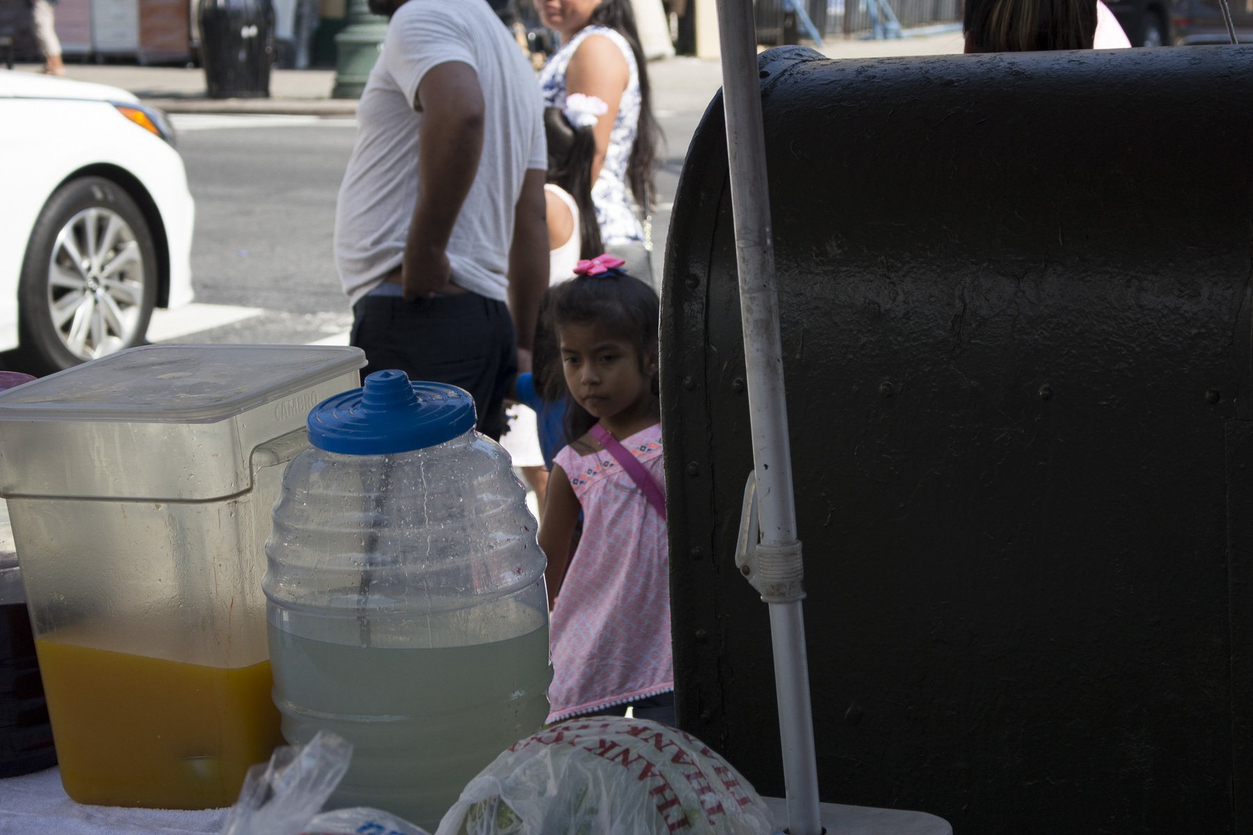  A little girl waiting with her mother to buy products from the fruit stand on the corner of 5th and 56th street. She is gazing at the limonada and jugo de mango that is for sale of August 22nd, 2017. 