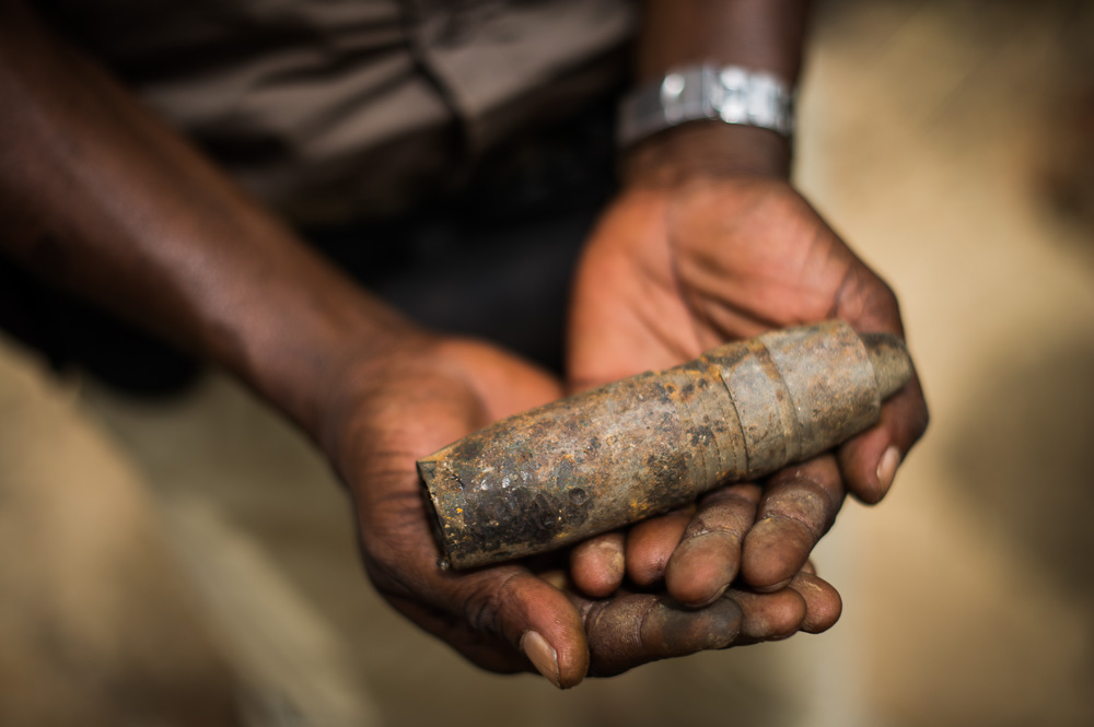  ​A de-mining worker holds an old munition round, lying on the ground in the countryside. 