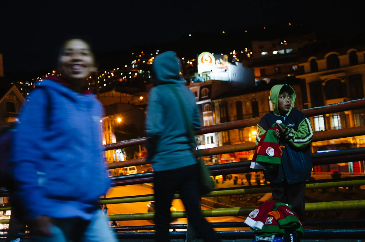 A young boy sells "Santa" hats on a pedestrian bridge at night in La Paz, Bolivia.