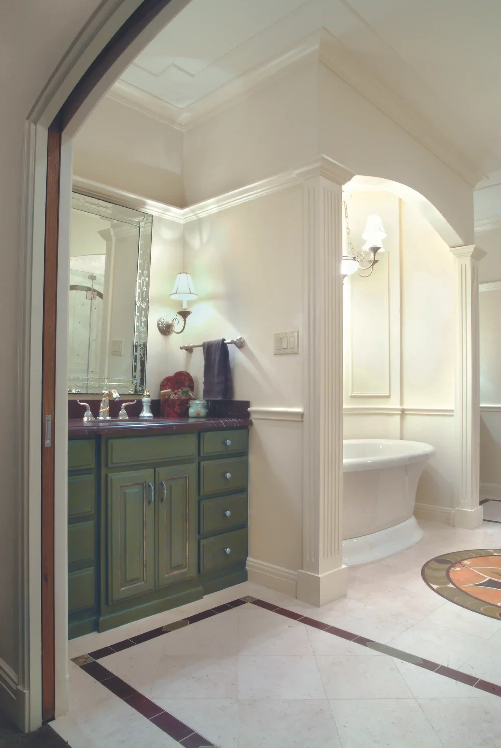 Elegant bathroom with green vanity, large mirror, wall-mounted lamps, a towel rack, and a soaking bathtub in a separate nook.