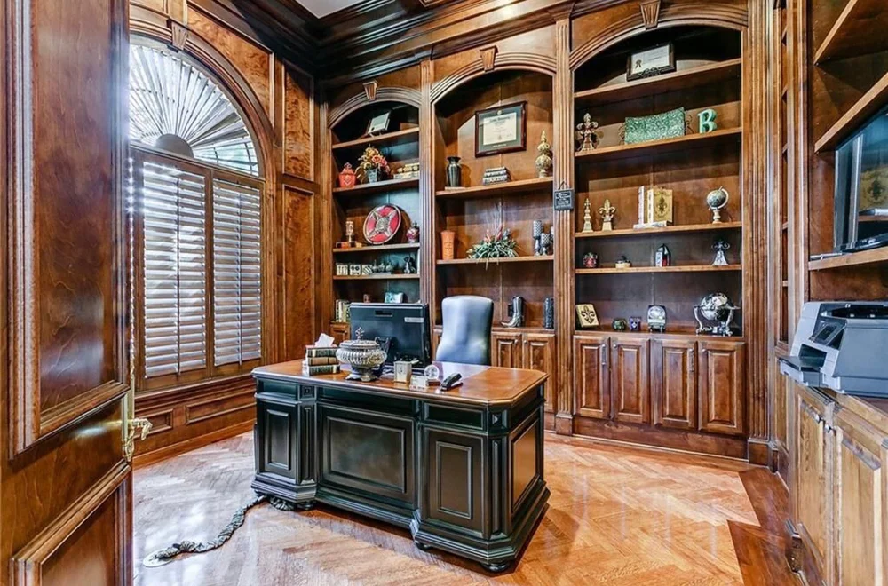 A luxurious office with wood-paneled walls, built-in bookshelves, a large window with shutters, and a central desk with a computer and decorative items.