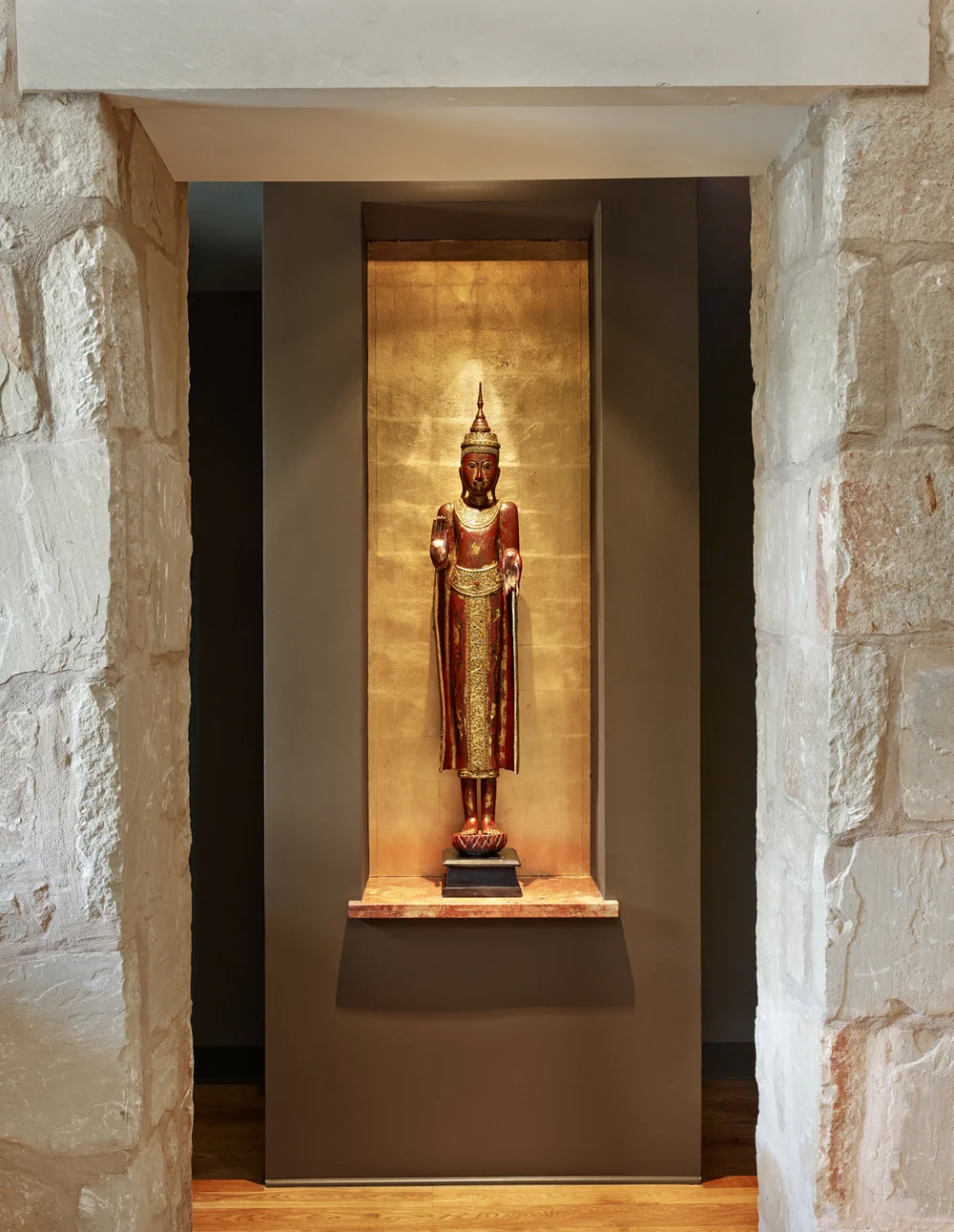 A traditional Thai-style wooden statue of a standing Buddha, decorated with gold and red accents, placed on a pedestal within a niche with a gold background.