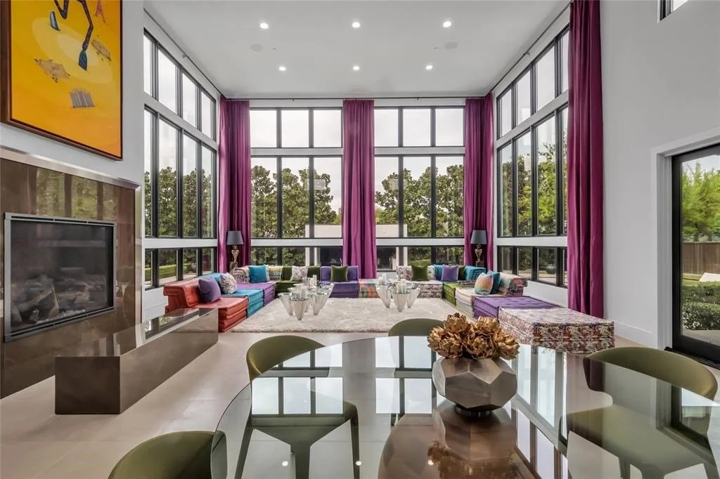 Bright living room with large windows, purple curtains, colorful sofas, and a fireplace, viewed from a dining area with green chairs and a decorative vase.