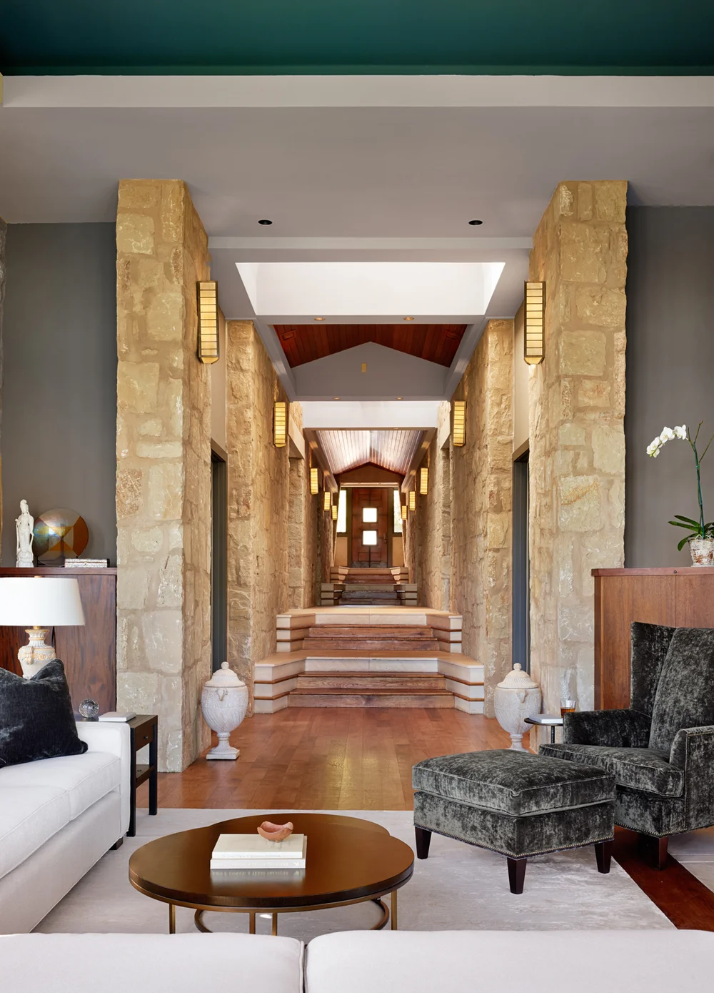 Interior view of a luxurious home with a stone and wood staircase leading to an upper level, framed by stone columns and wall sconces; modern seating area in the foreground with a white sofa, black armchair, and a round coffee table.
