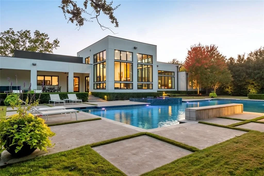 Modern house with large glass windows, surrounding a swimming pool, with outdoor patio furniture, greenery, and trees in the background, during sunset.