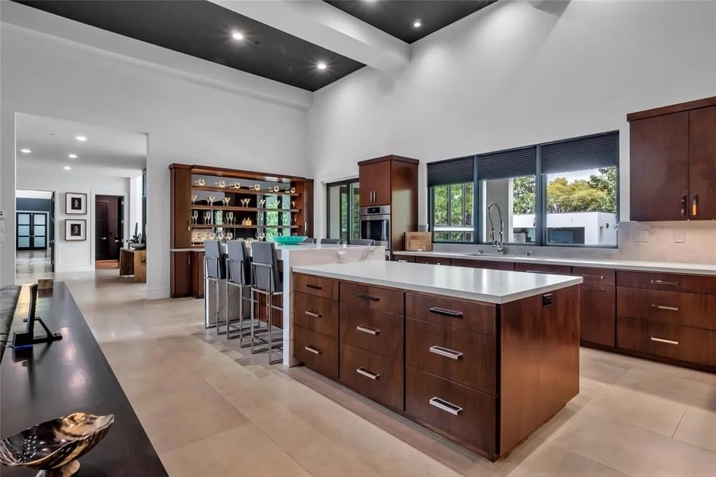 Modern kitchen with dark wood cabinets, white countertops, stainless steel appliances, and large windows showing outdoor greenery.