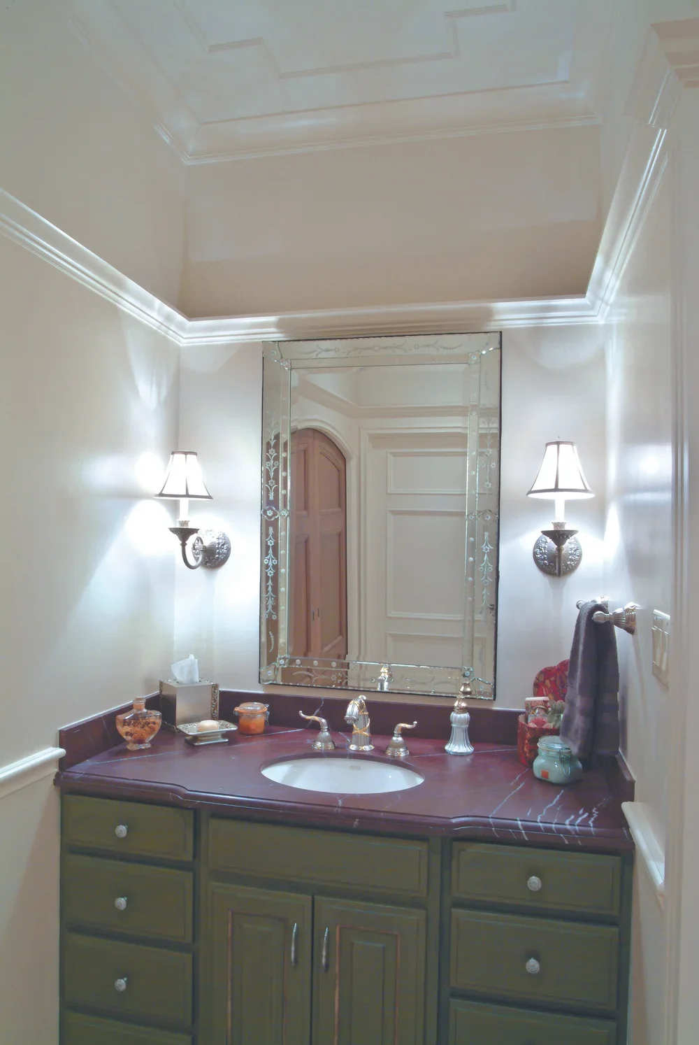 A bathroom vanity with a dark countertop, green cabinets, a mirror with a decorative border, and two wall-mounted lights.