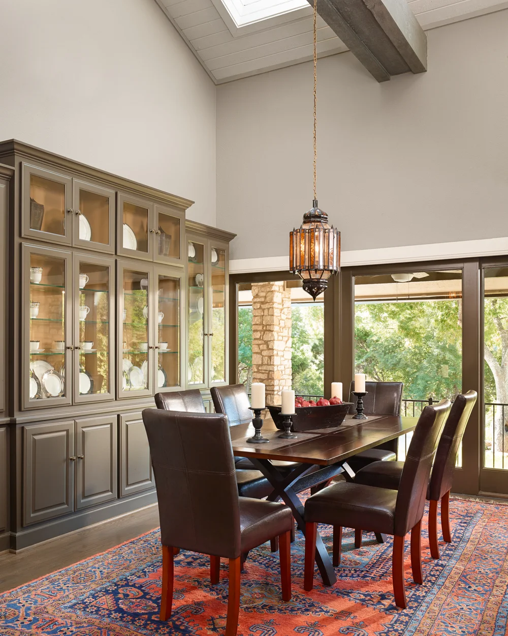 A dining room with a wooden table surrounded by six leather chairs, a large built-in china cabinet with glass doors displaying dishes, a hanging pendant light, and sliding glass doors leading to an outdoor area with trees.