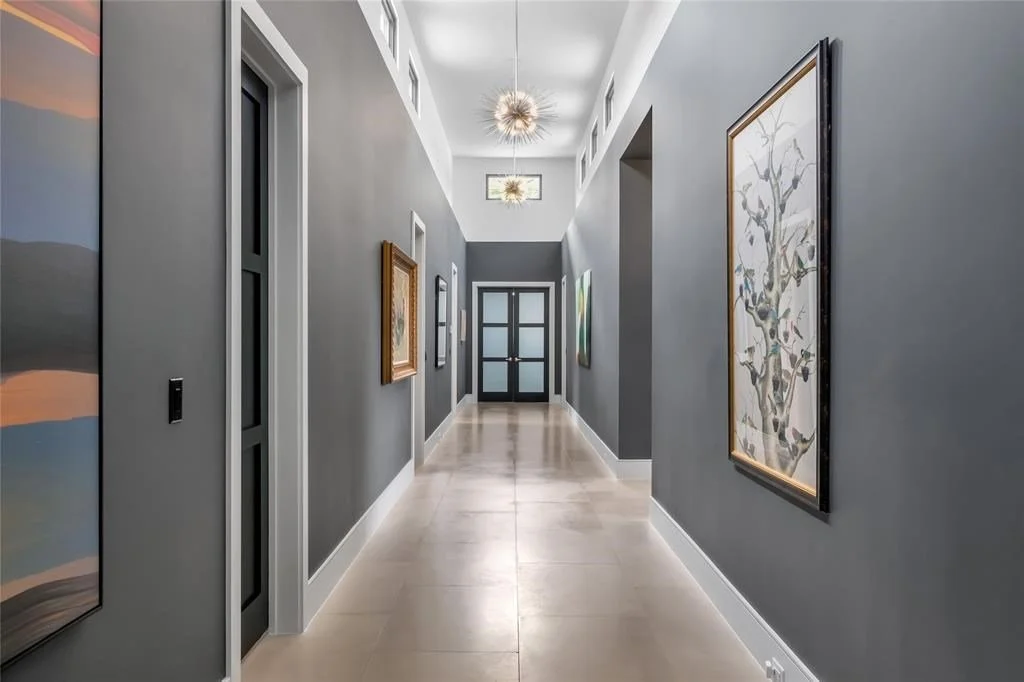 Interior hallway with gray walls, framed artwork, modern ceiling light fixtures, and glass-paneled front door at the end.