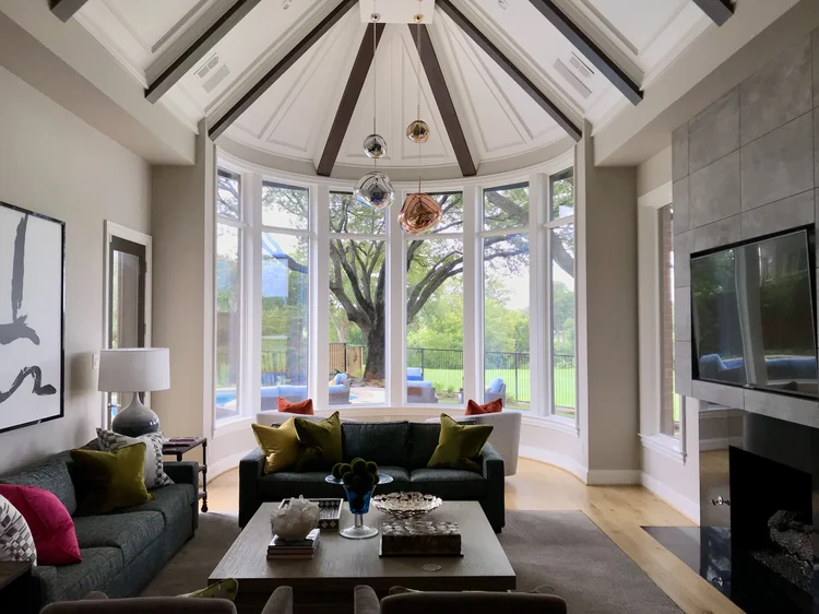 Living room with large bay window overlooking a backyard with a tree. Modern furniture, including a dark sofa with colorful pillows, a coffee table with decorative items, and a wall-mounted flat-screen TV. Artistic pendant lights hanging from the cei