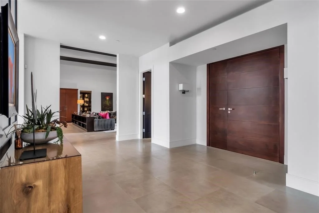 Modern interior with white walls, beige tile flooring, and wooden accents, including a large wooden door and a console table with plants.