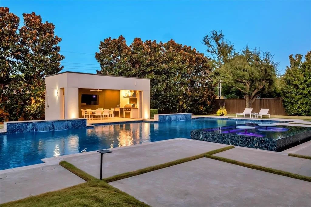Backyard with swimming pool, hot tub with illuminated jets, poolside lounge chairs, outdoor kitchen and dining area, lush trees, and a clear evening sky.