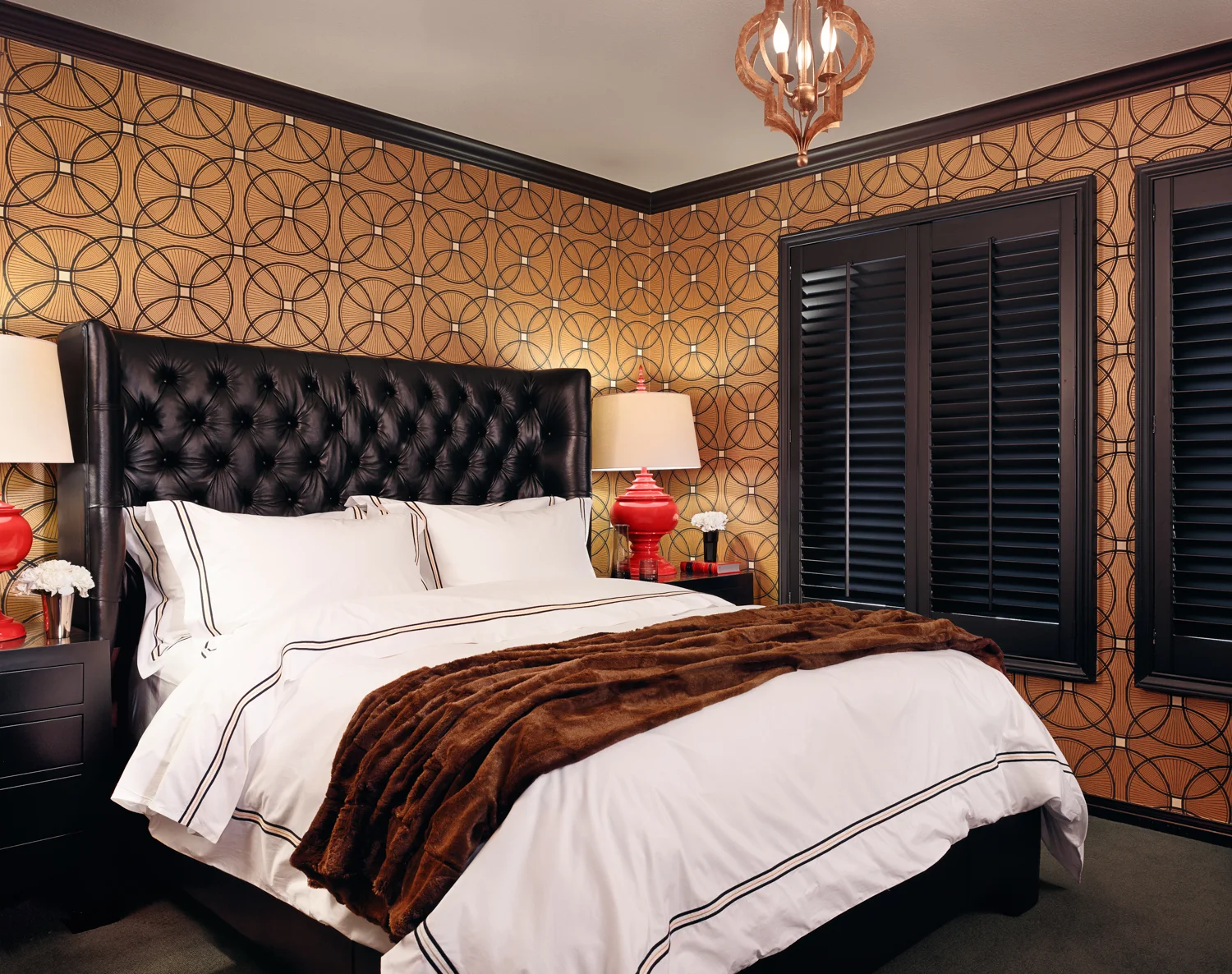A bedroom with a large bed featuring a tufted black leather headboard, white bedding with black trim, and a brown fur throw. Two black nightstands with red and white lamps, a decorative wall with a geometric pattern, and black louvered shutters on th