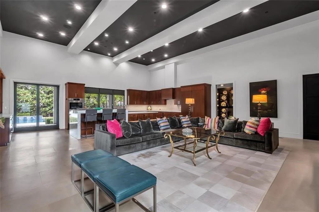 Spacious living room with high vaulted ceiling, dark wood beams, and modern recessed lighting. Features a large L-shaped sofa with colorful pillows, a glass coffee table with gold legs, and a blue bench. Open kitchen with wooden cabinets, bar stools,