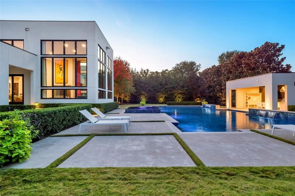 Modern house with large glass windows, outdoor pool with surrounding lounge chairs, landscaped yard, trees in the background, and a covered outdoor living space at sunset.